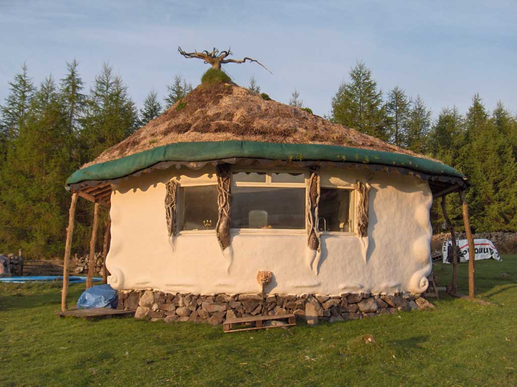 Ronde rieten hut met kleine ramen op een grasveld, met bomen op de achtergrond.