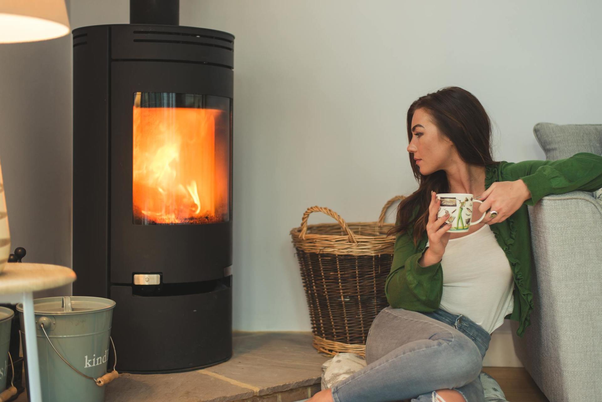 girl with cup of coffee looking at cosy wood burner lit with flames