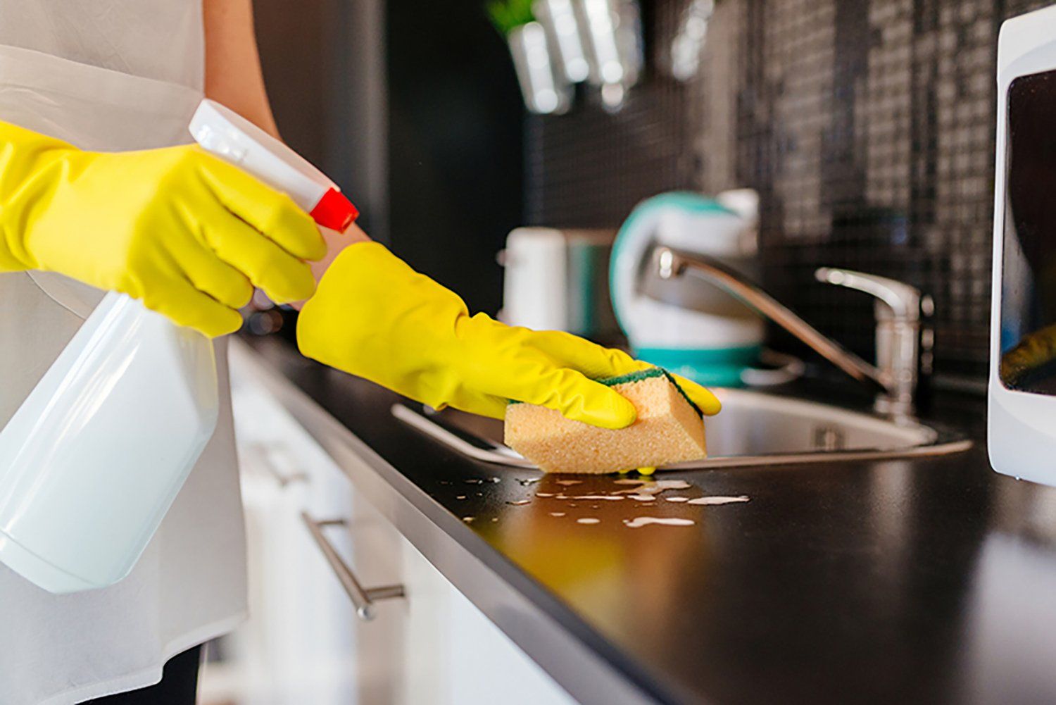 Person wearing yellow gloves spraying and wiping a kitchen countertop with a sponge.