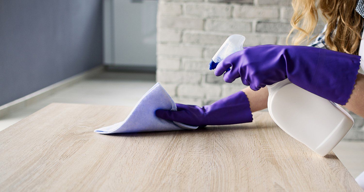 Person in purple gloves spraying cleaner onto a wooden table and wiping with a purple cloth