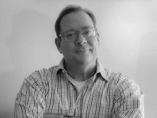 A man wearing glasses and a plaid shirt is smiling in a black and white photo.