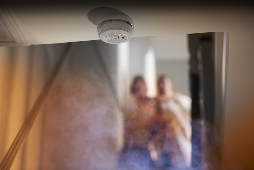 smoke rises up towards a second floor in a two two storey house . Upstairs we can see a parent carrying a small child and running from a bedroom as they have been woken by the smoke alarm which is shown in focus at the edge of the ceiling leading to the stairwell