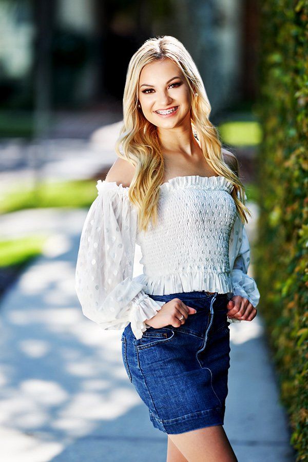 A woman in a white off the shoulder top and denim skirt is standing on a sidewalk.