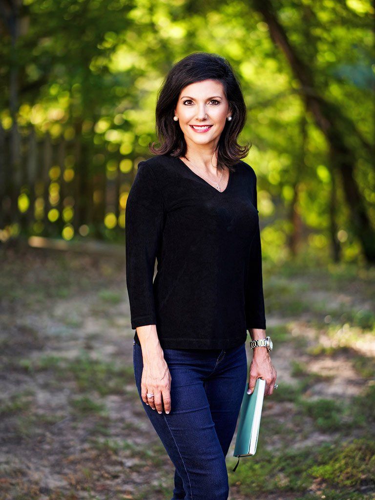 A woman in a black shirt and jeans is standing in the woods holding a book.