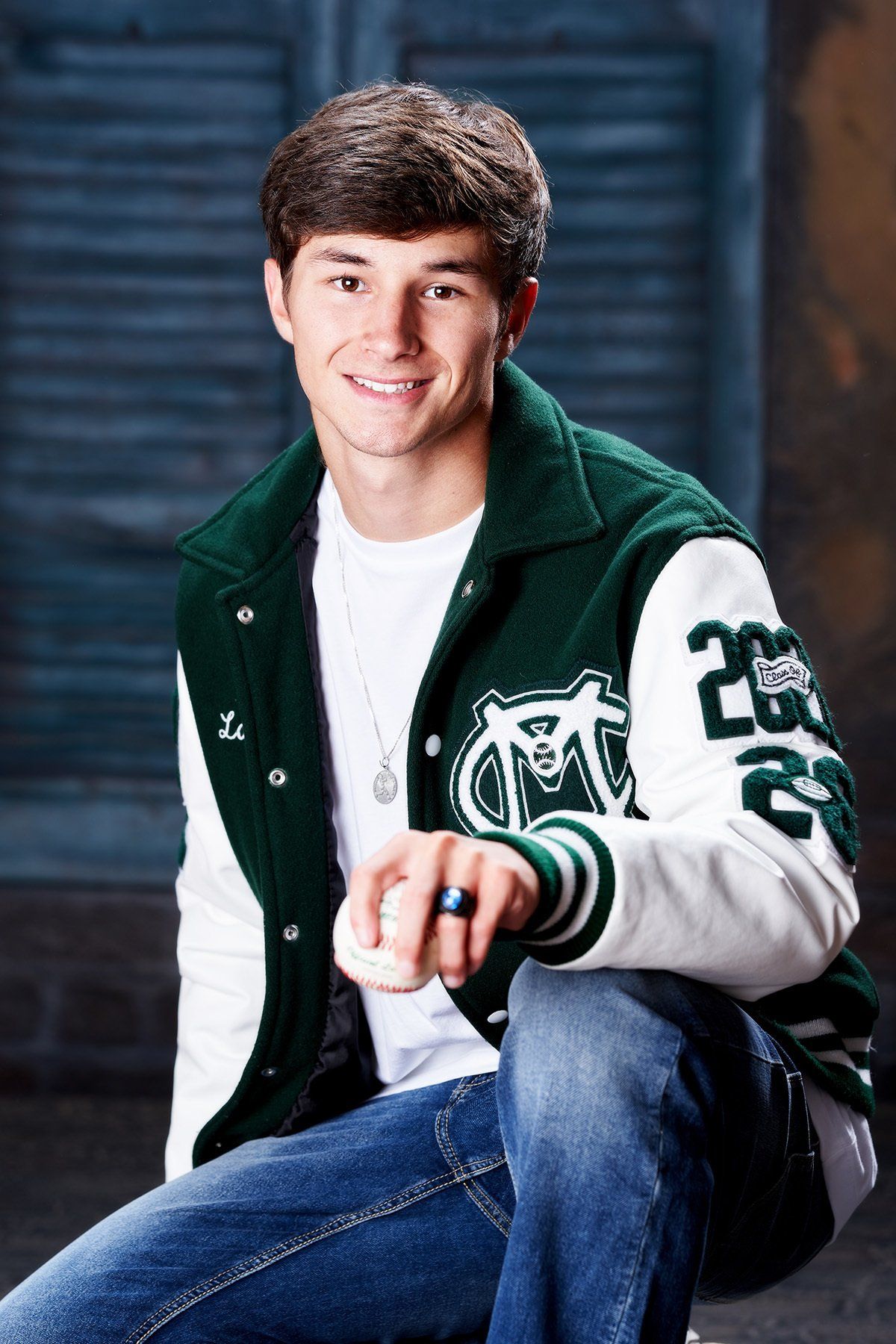 A young man wearing a green and white varsity jacket is kneeling down holding a baseball.