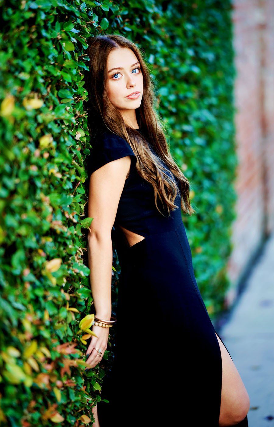 A woman in a black dress is leaning against a green hedge.