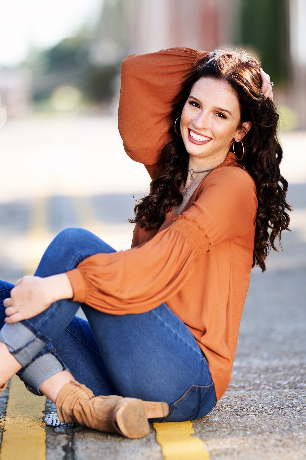 A woman is sitting on the sidewalk with her legs crossed and smiling.