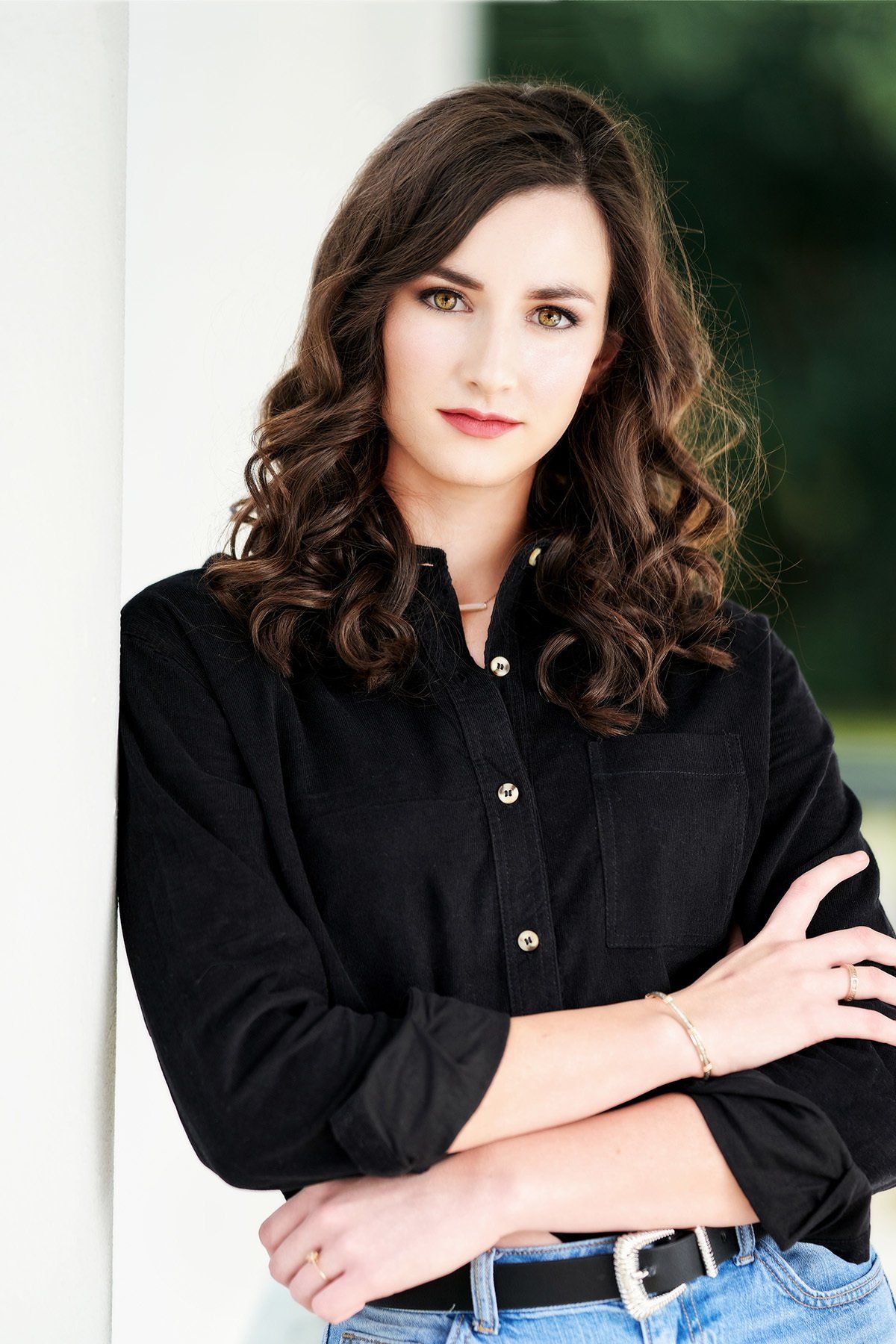 A woman in a black shirt and jeans is leaning against a wall with her arms crossed.