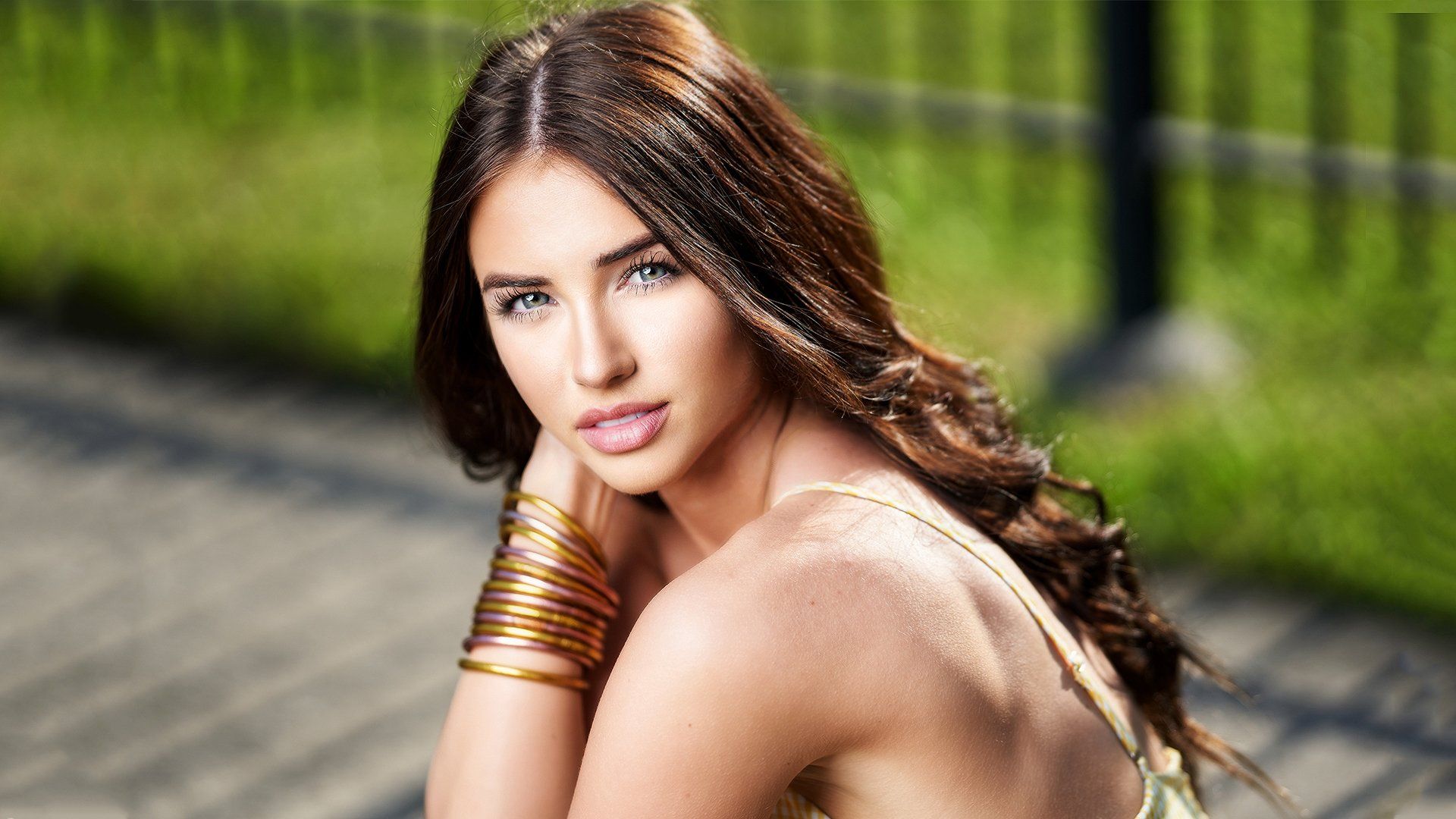 A woman with long hair and bracelets on her wrists is sitting on a sidewalk.