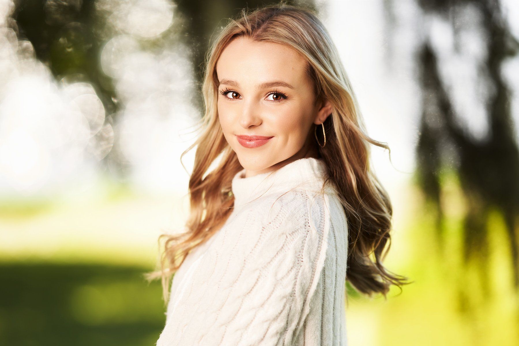 A young woman in a white sweater is standing in a park.