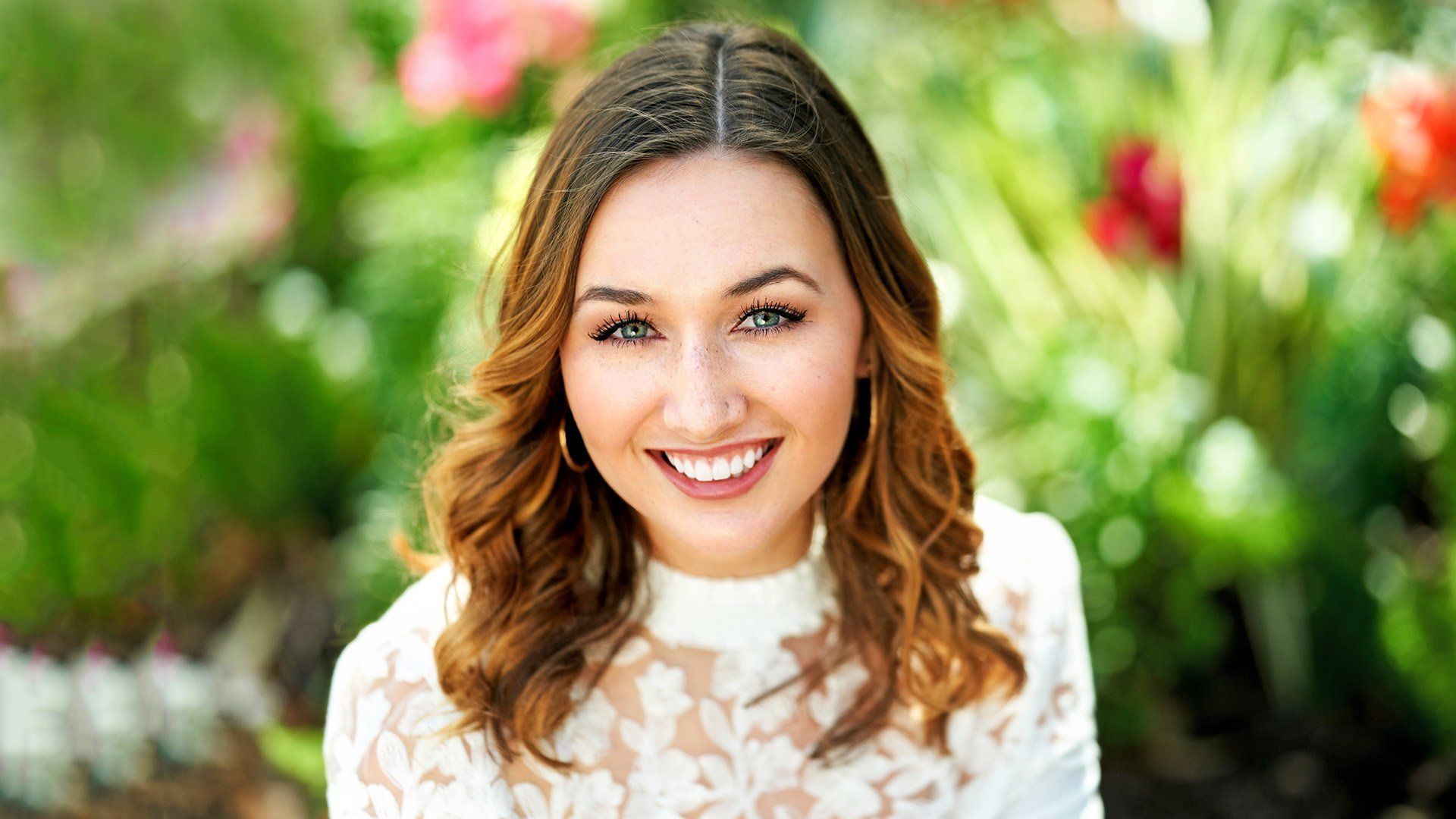 A woman in a white lace top is smiling in front of flowers.