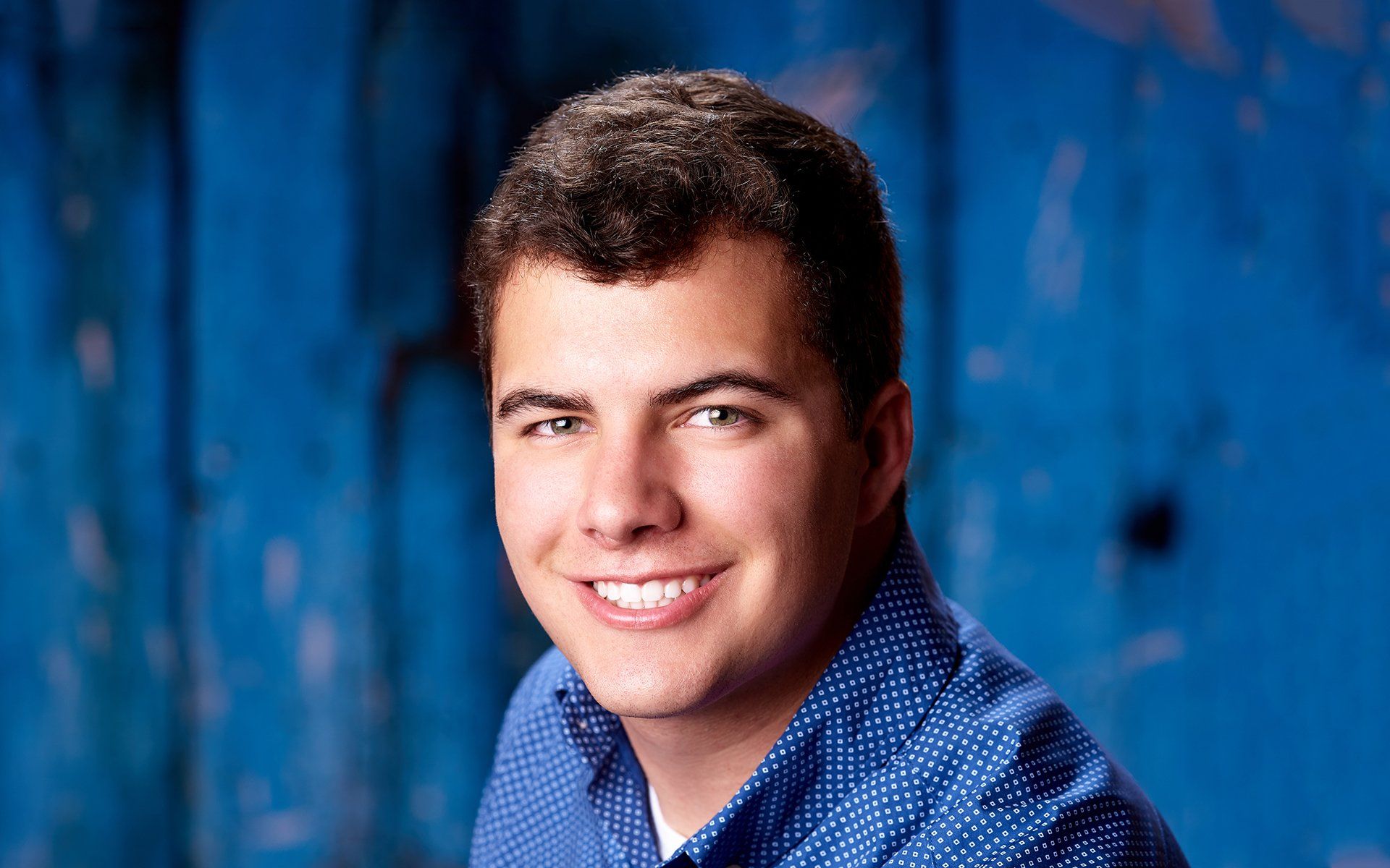 A young man in a blue shirt is smiling in front of a blue wall.