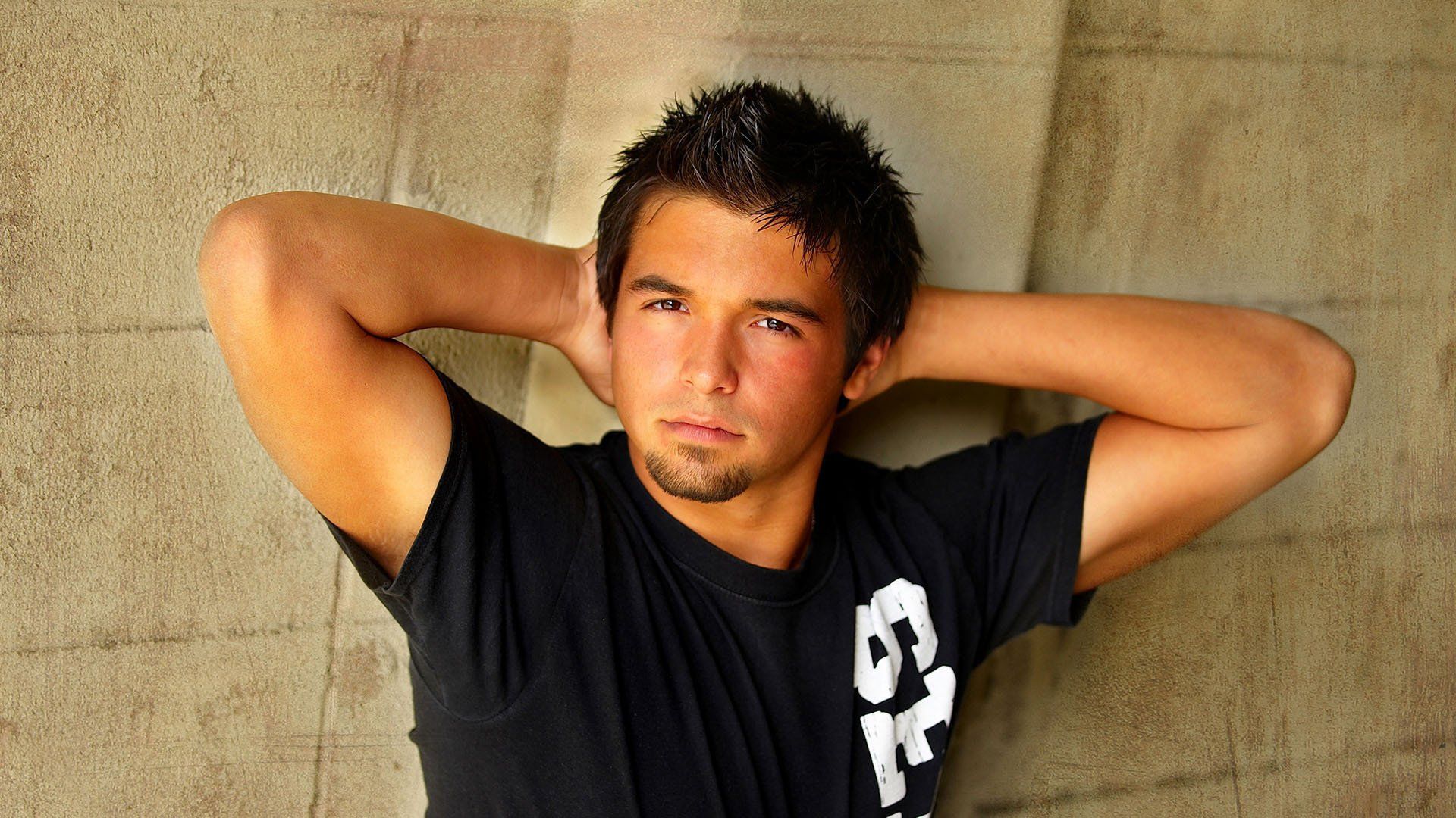 A young man with his hands behind his head is wearing a black t-shirt with the letter g on it
