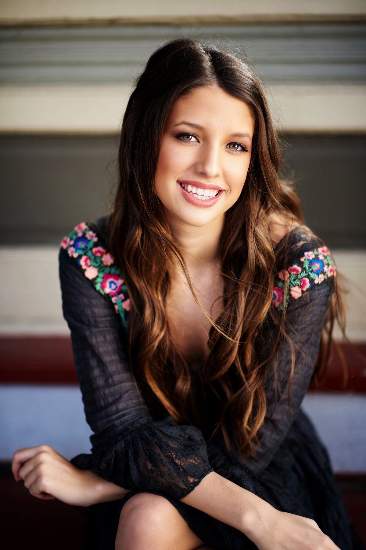 A woman in a black dress is sitting on a bench with her legs crossed and smiling.