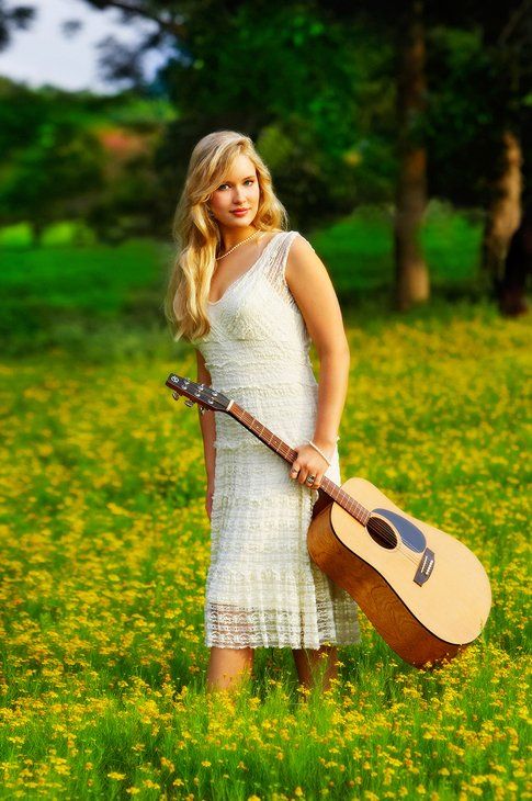 A woman in a white dress is holding a guitar in a field of yellow flowers.