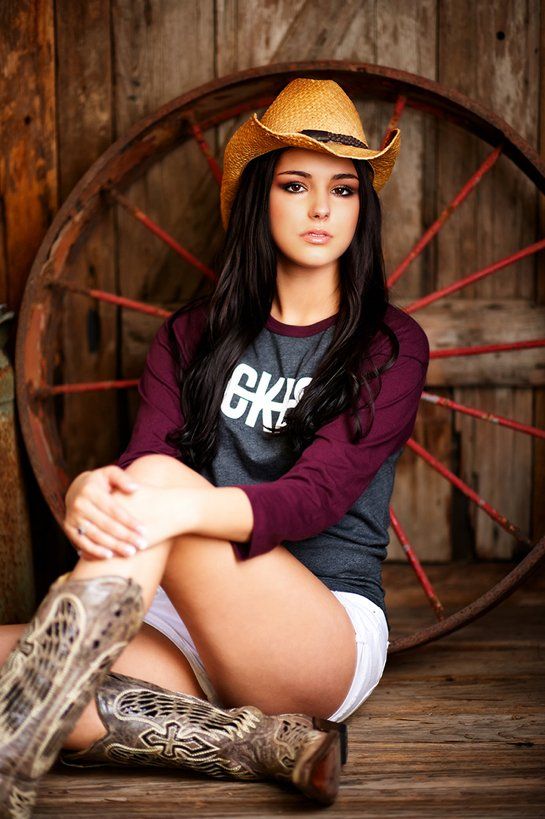 A woman wearing a cowboy hat and boots is sitting next to a wagon wheel.
