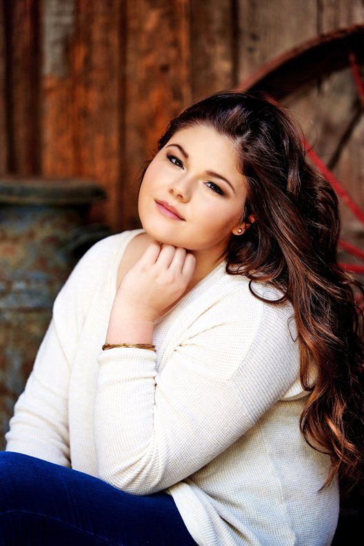 A young woman in a white sweater and blue jeans is sitting in front of a wooden wall.