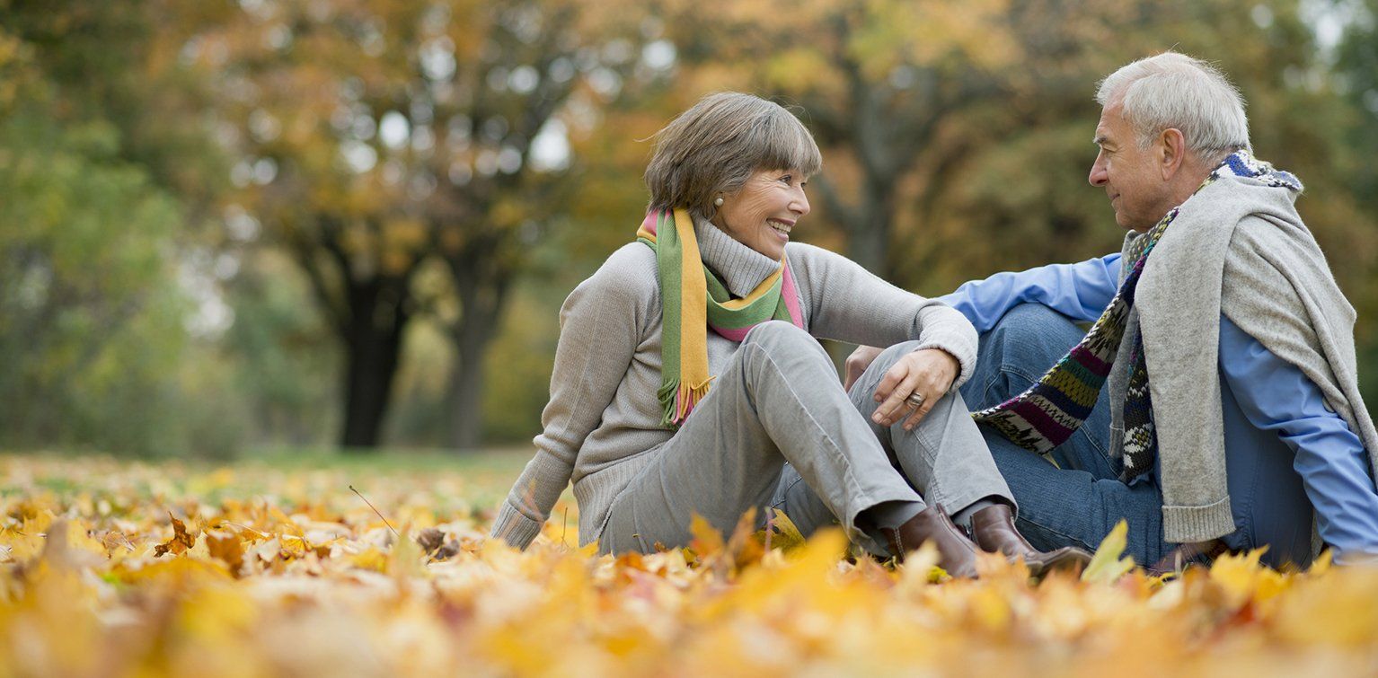 An elderly couple is sitting in a pile of leaves in a park.