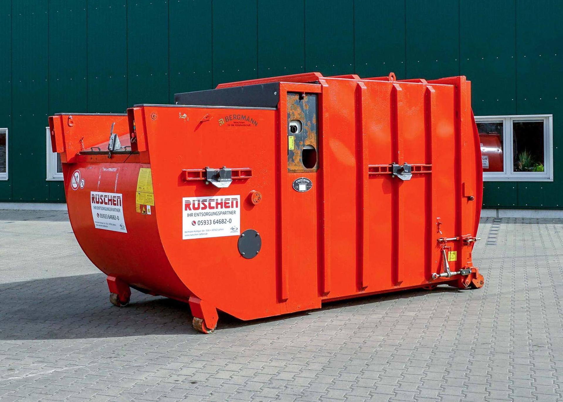 Rüschen Selbstpressbehälter Vor einem grünen Gebäude steht ein großer orangefarbener Selbstpressbehälter.