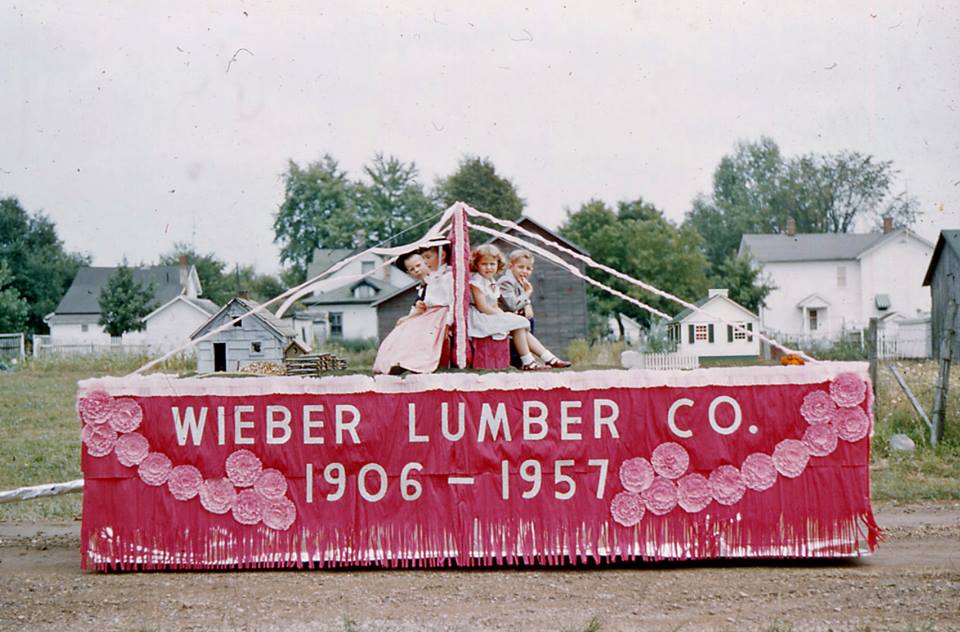 A red parade float for Wieber Lumber Co. 1906–1957 decorated with flowers, featuring three children sitting on top.