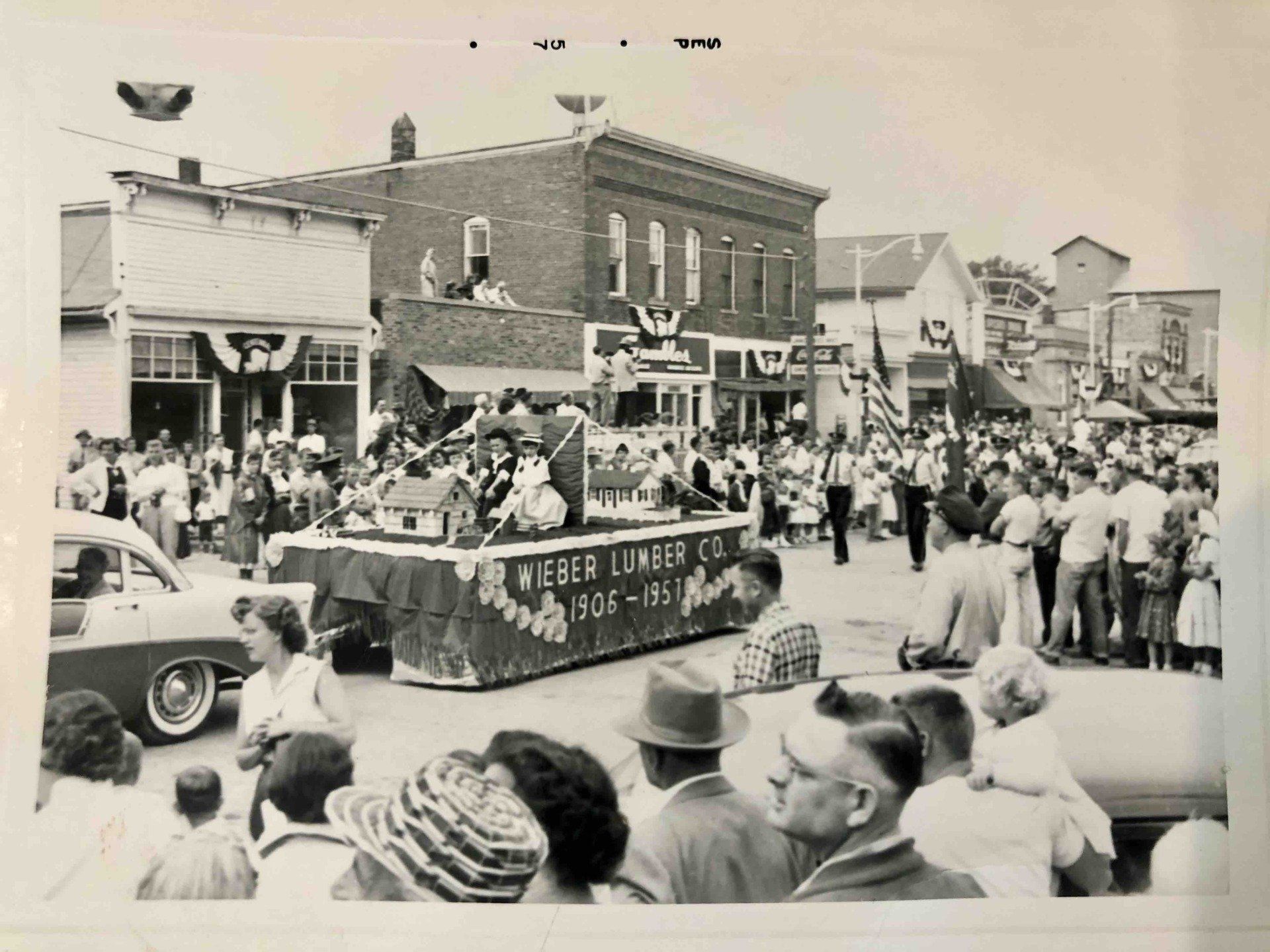 A black-and-white parade scene featuring a Wieber Lumber Co. float with 1906-1956 written on the side and a crowd gathering.