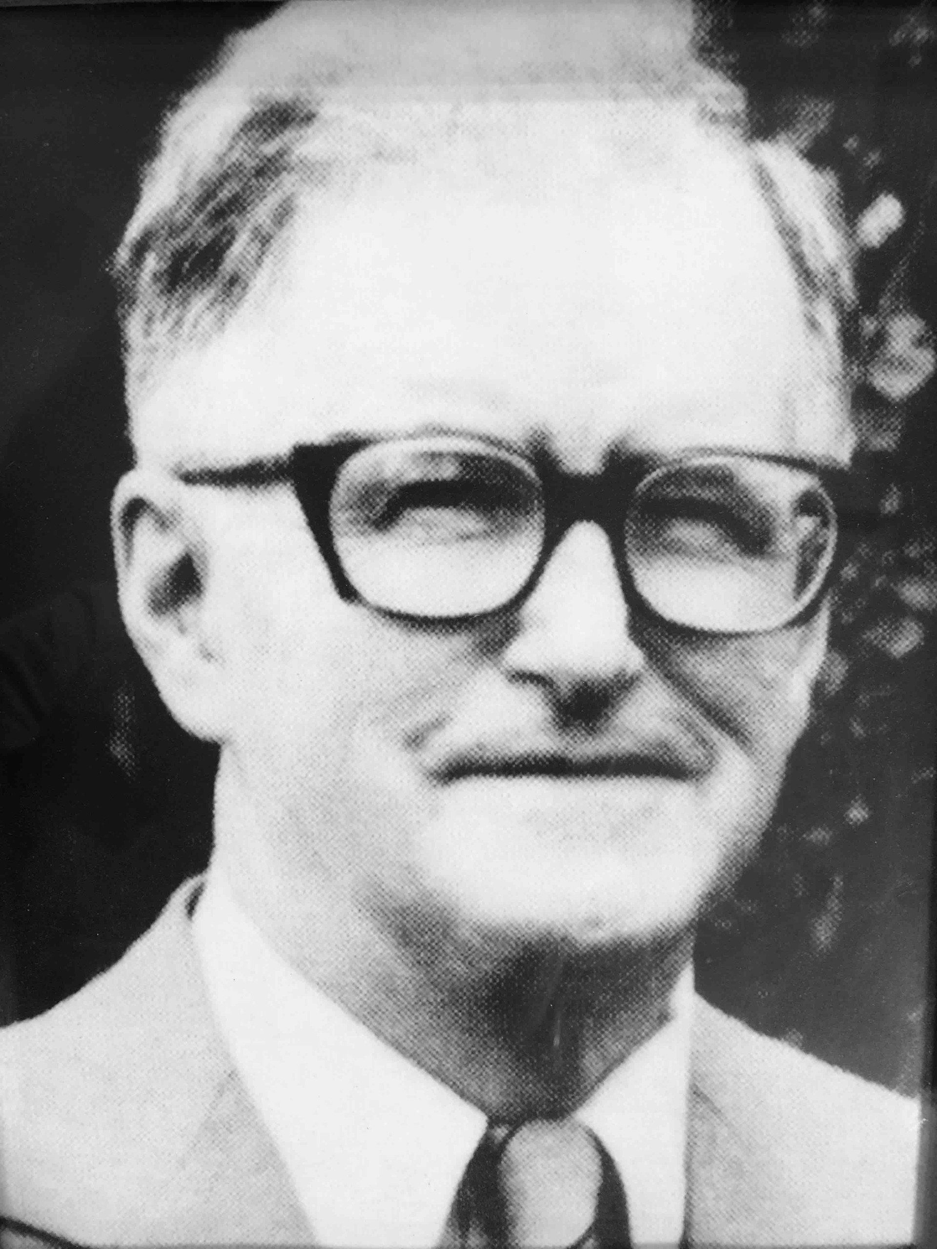 A portrait of Sir William Hayter wearing thick-rimmed glasses, a suit, and a tie.