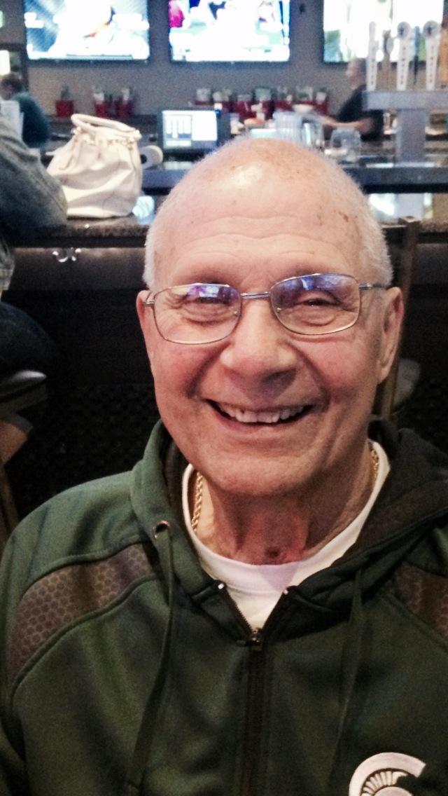 A smiling person wearing glasses and a green zip-up jacket at a bar or restaurant with television screens in the background.