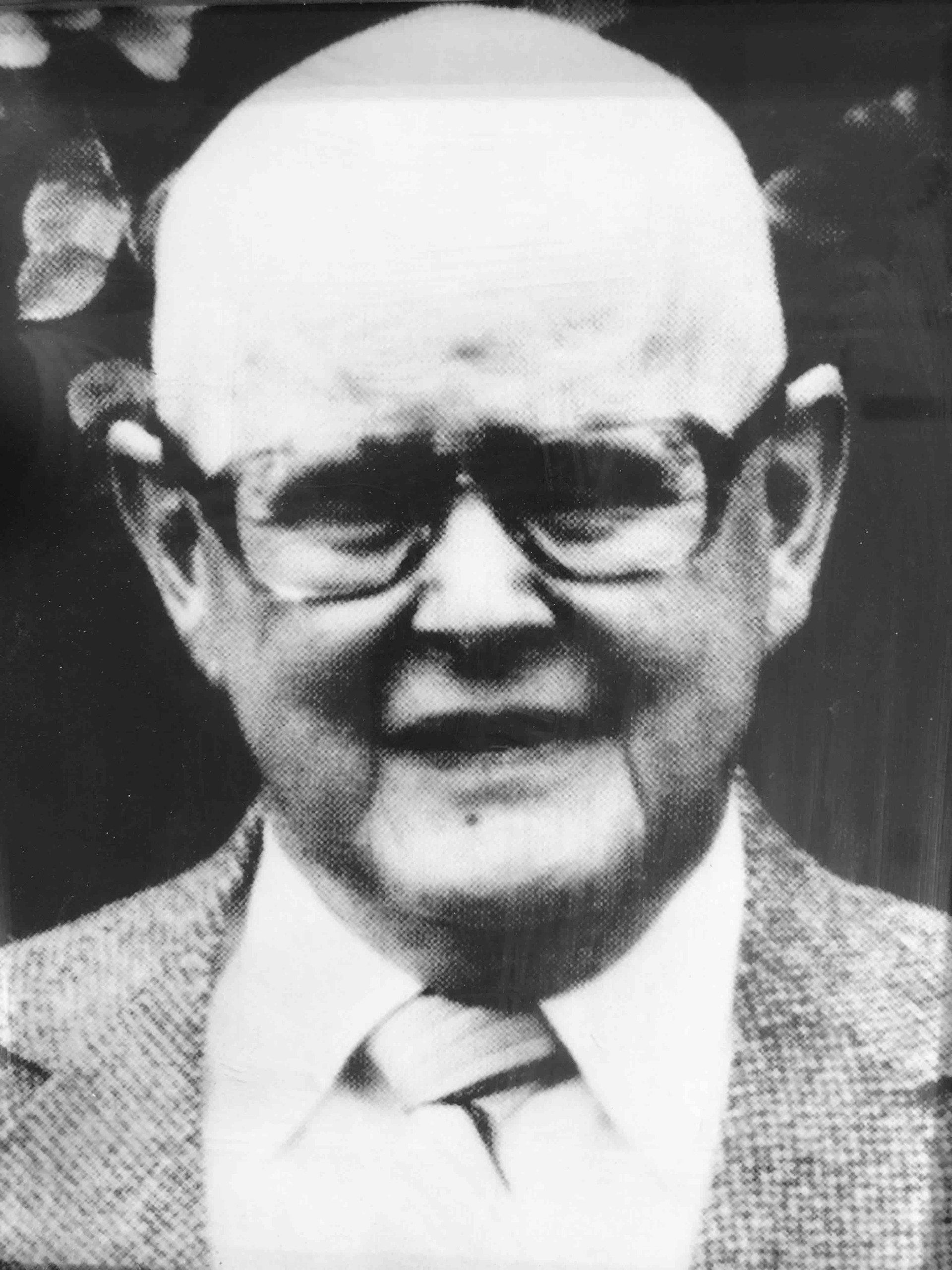 A portrait of a person with a shaved head wearing glasses, a collared shirt, and a blazer.