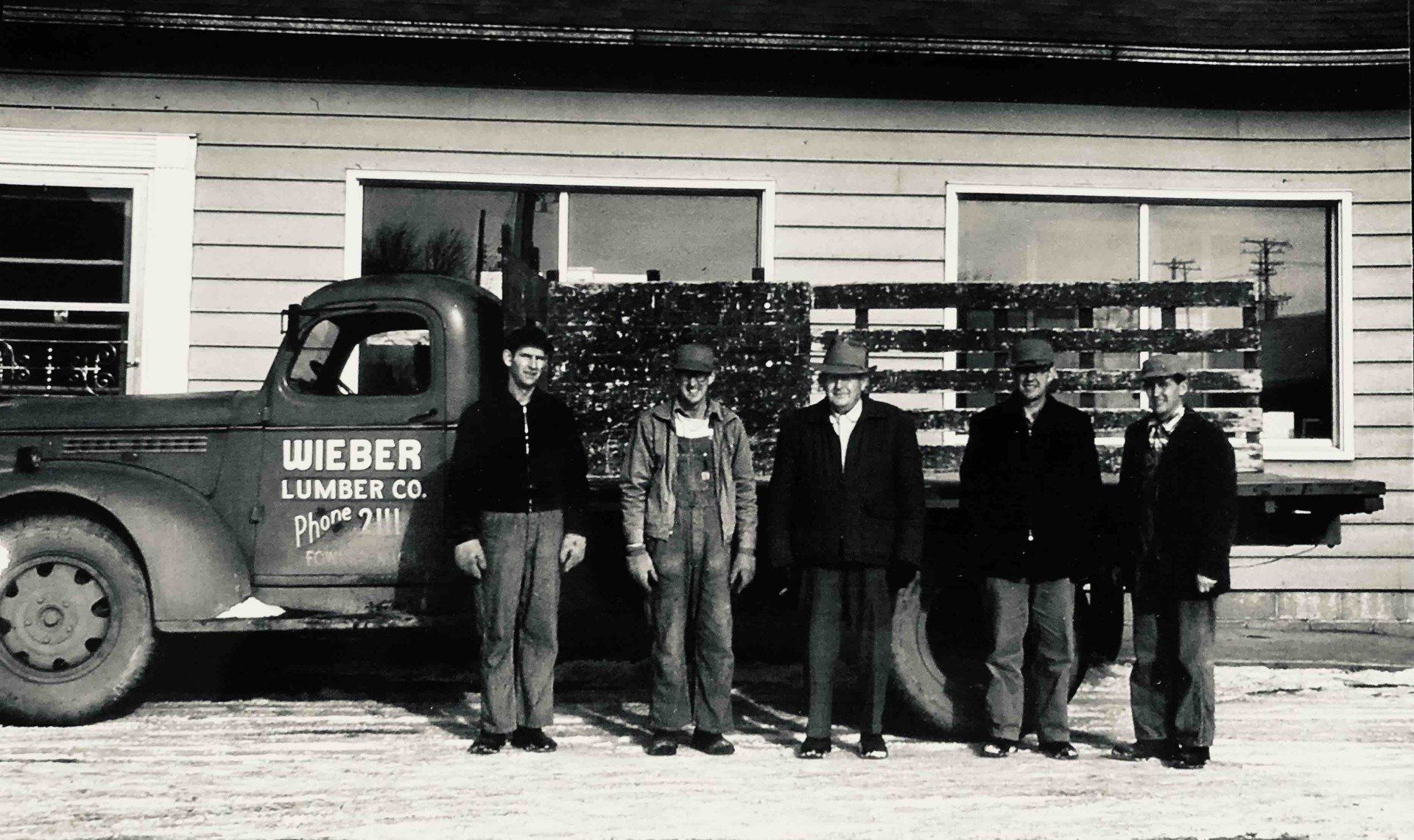 Five men stand in front of a Wieber Lumber Co. delivery truck parked outside a building.