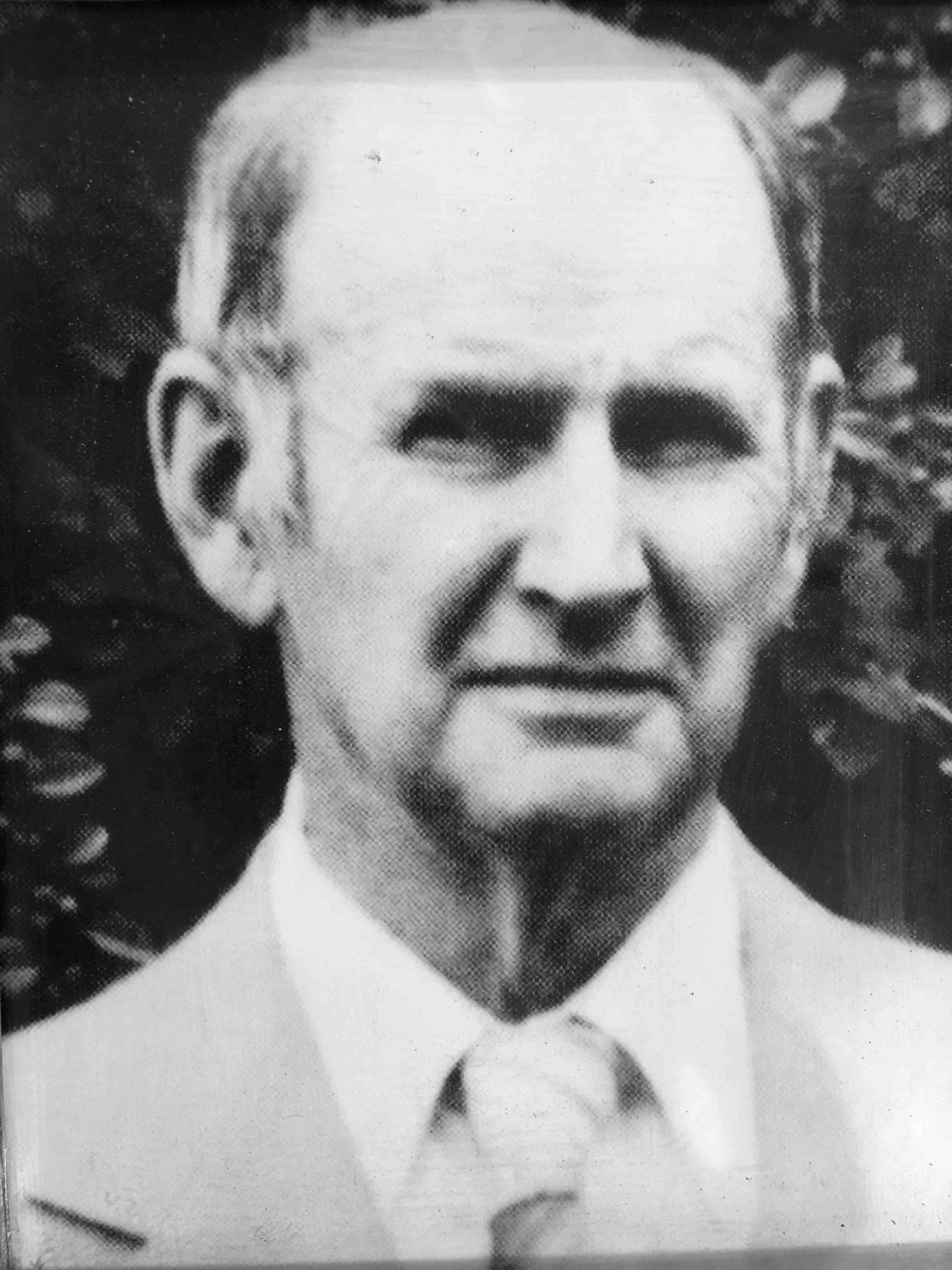 A head-and-shoulders portrait of an individual wearing a collared shirt and tie, set against a leafy, dark background.