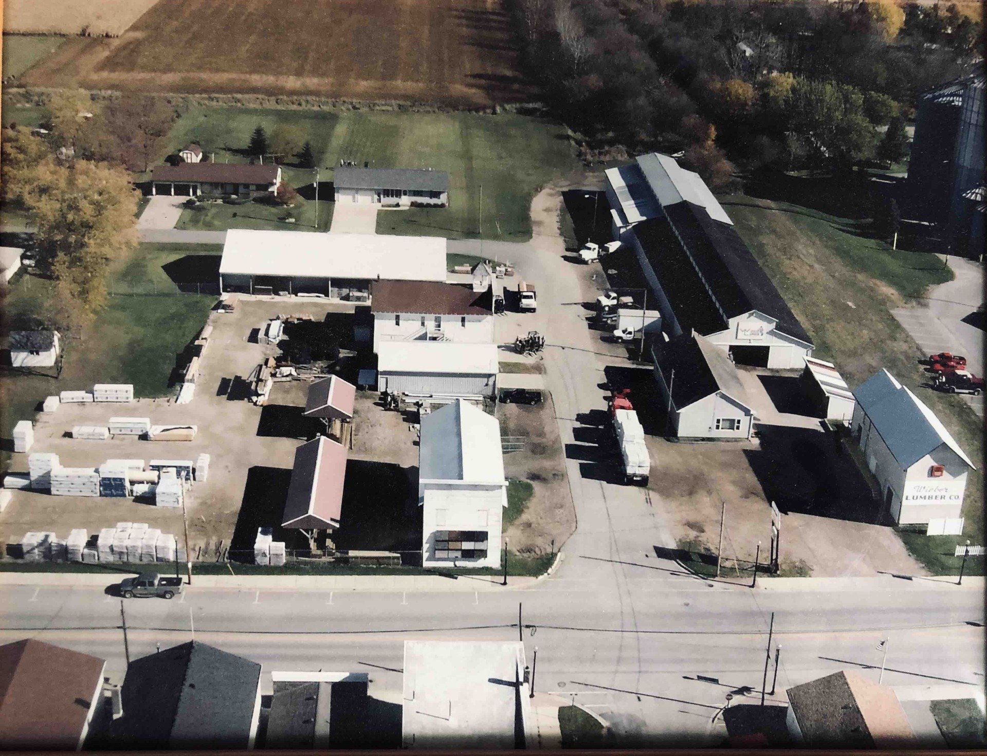 An aerial view of a rural business complex with multiple buildings, storage areas, and vehicles, set near a road.
