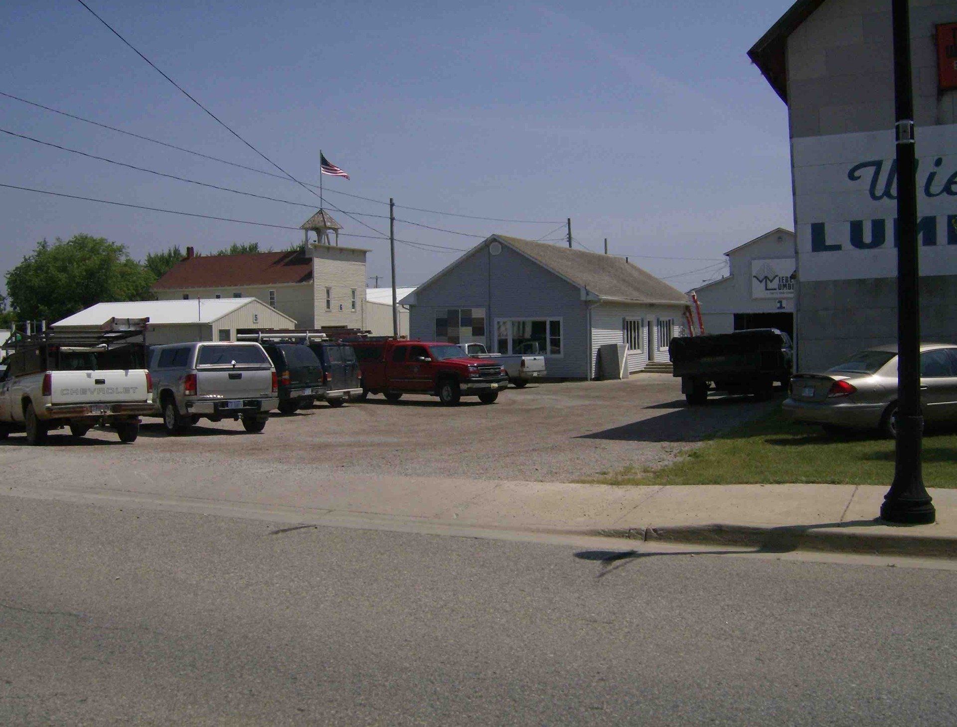 A gravel parking lot in front of commercial buildings and a lumber yard under a clear blue sky.