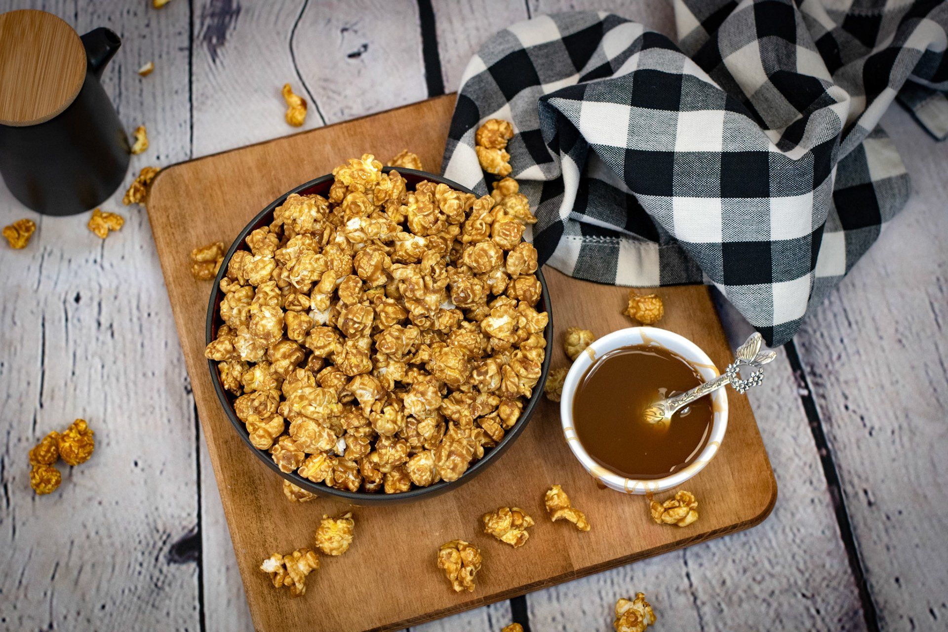 A bowl of caramel popcorn and a cup of caramel sauce on a wooden cutting board.