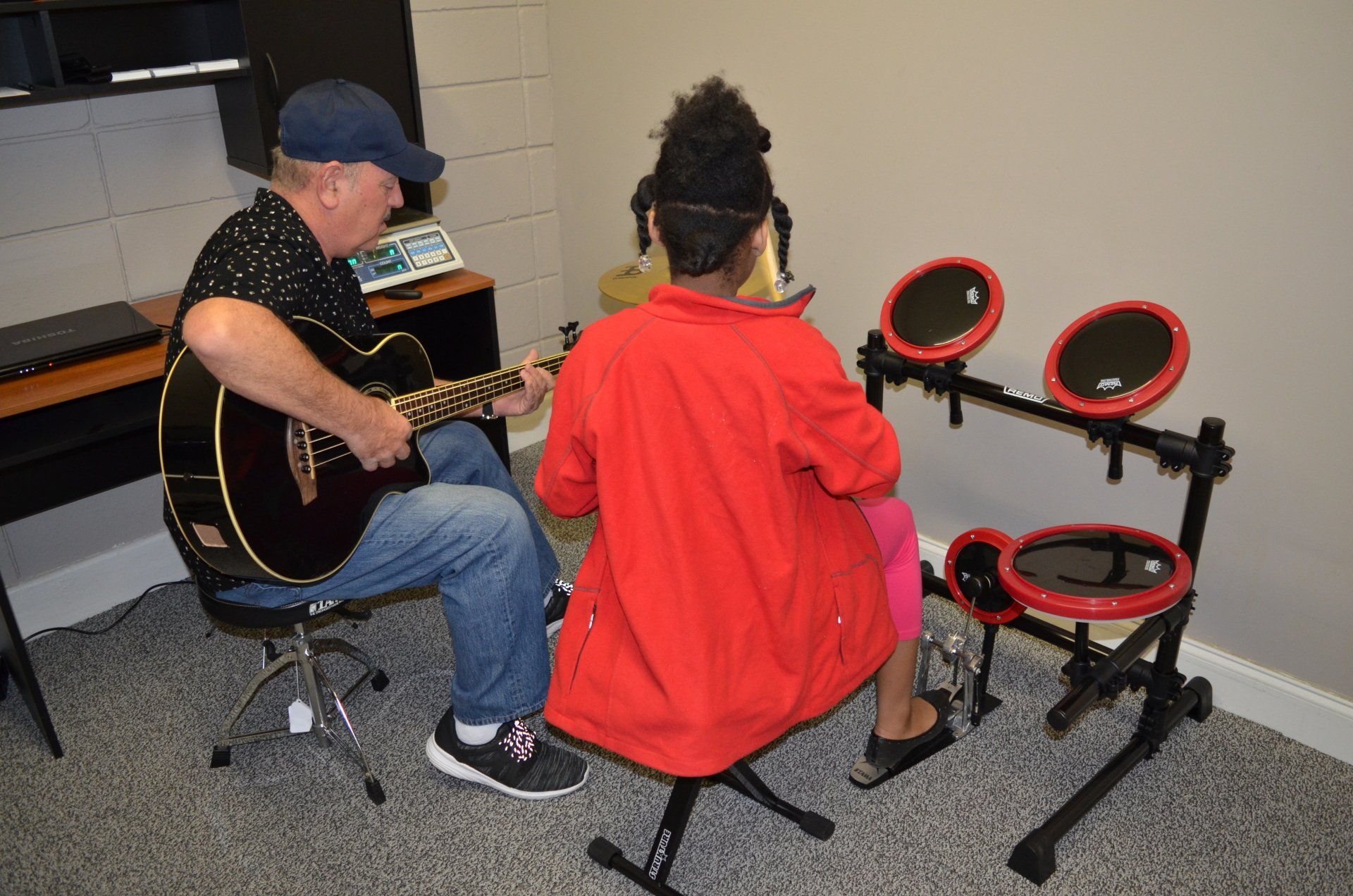 Man playing guitar and person drumming electronic drums in a music room.