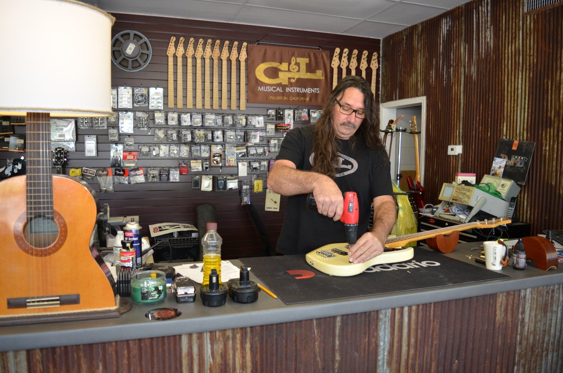 Man using a drill on an electric guitar at a repair shop counter with a classical guitar to the left.