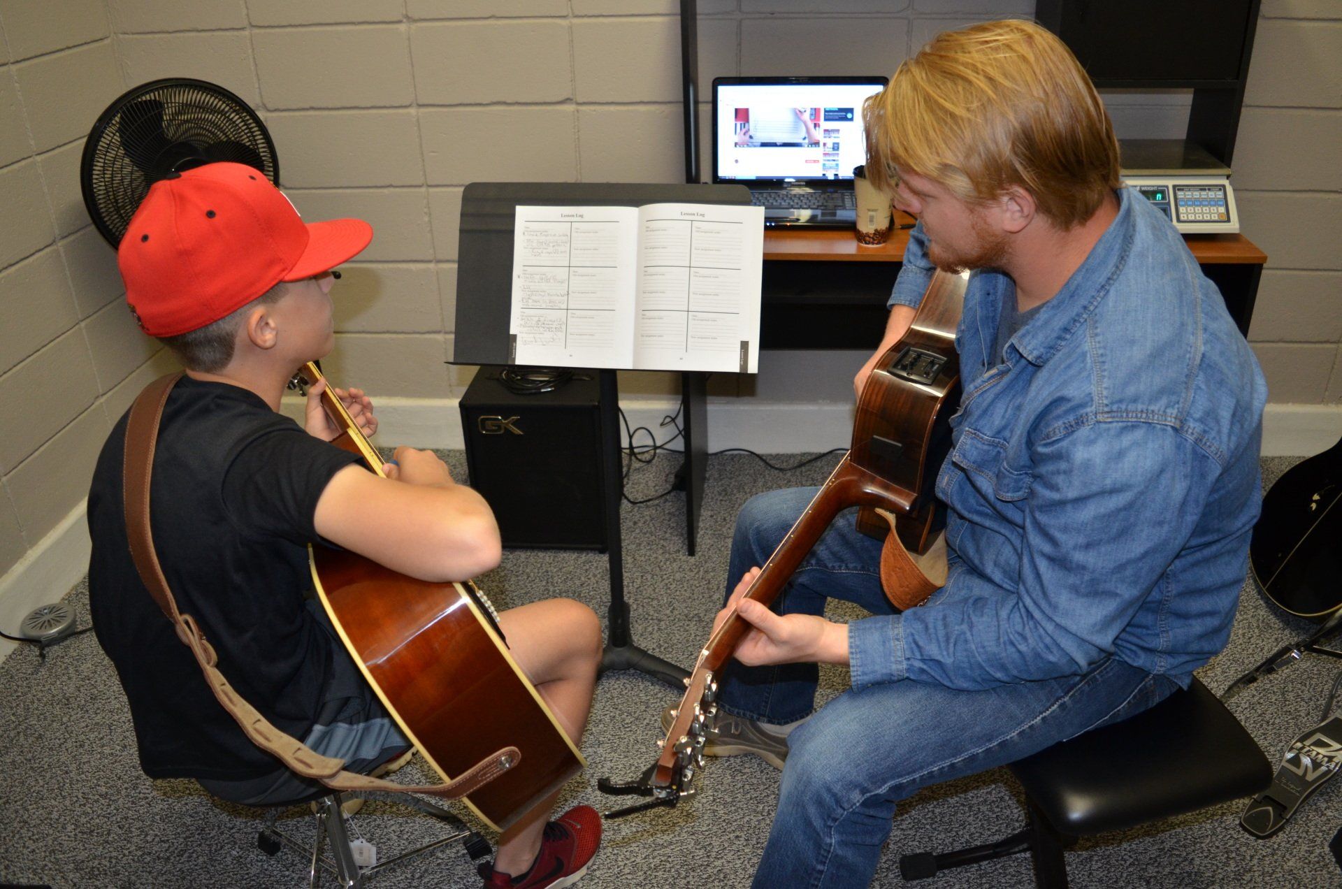 Man teaching a student guitar. They're in a music studio, studying sheet music, and there's a computer in the background.