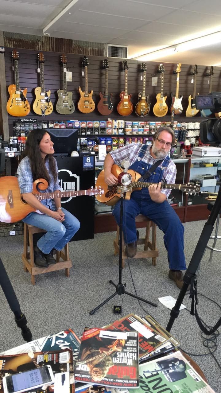 Two people in a music store, playing guitars. One sings; the other plays with overalls. Guitars on the wall.
