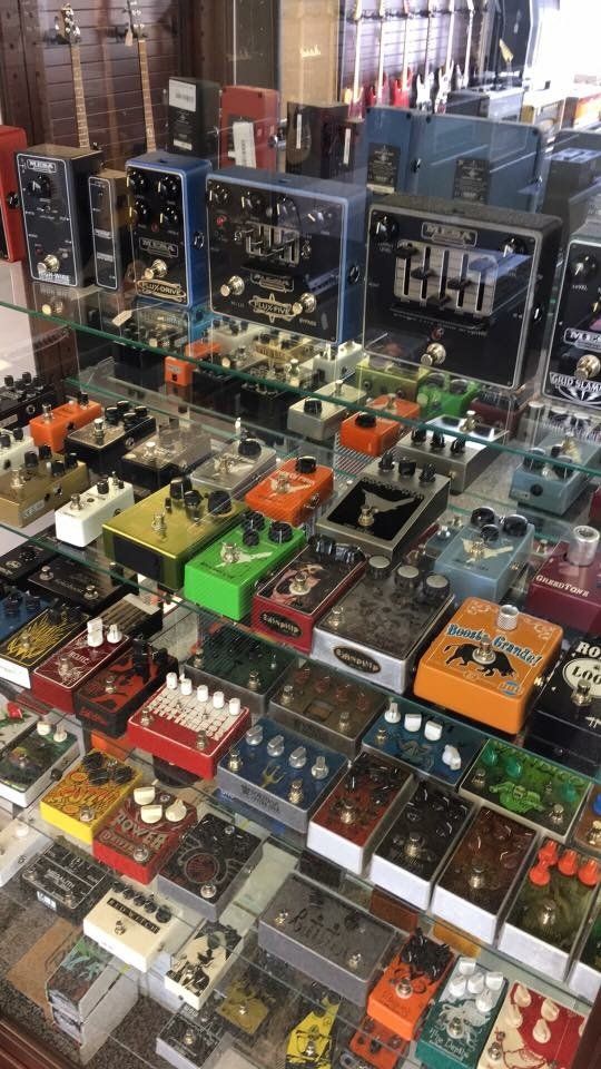 Guitar effects pedals in a display case, various colors and styles, with amplifiers in the background.