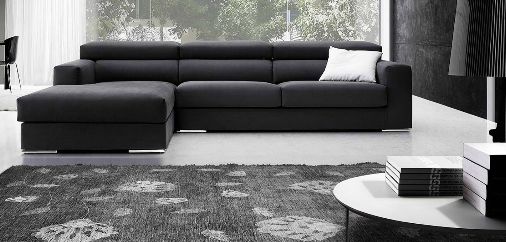 Soggiorno moderno con divano angolare nero, cuscino decorativo bianco, tappeto e tavolino rotondo vicino alle grandi finestre.