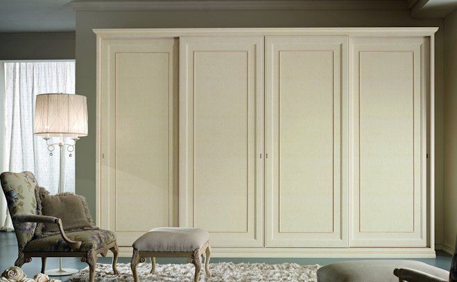 Armadio a muro color crema con ante a pannelli in una stanza luminosa, accanto a una poltrona, un pouf e una lampada da terra.