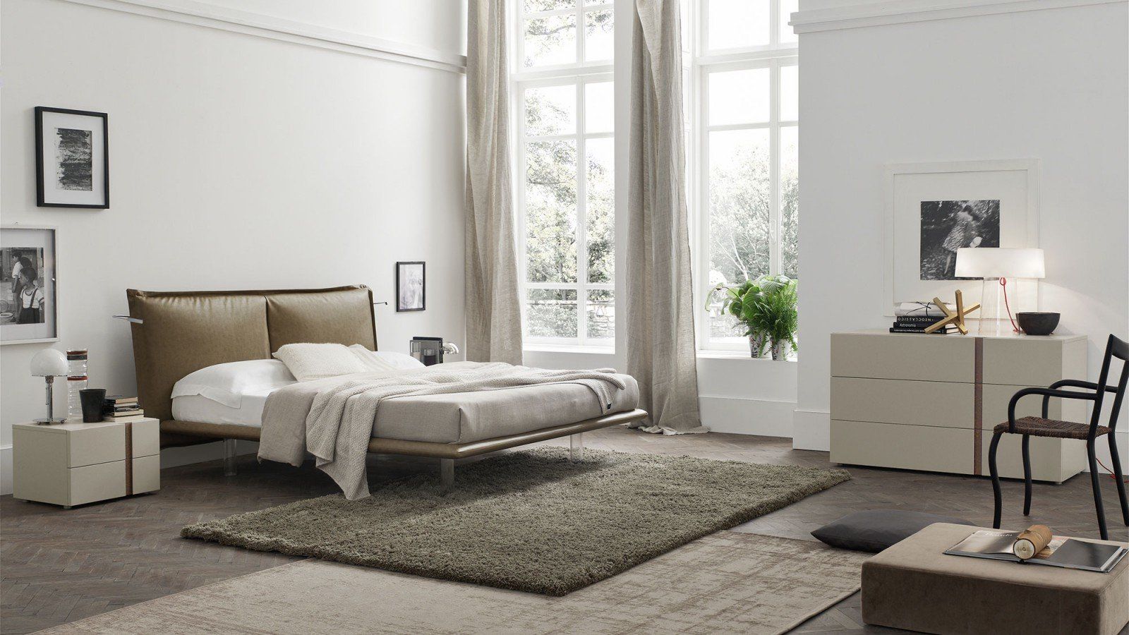 Camera da letto minimalista con letto, tappeto, scrivania, sedia e ampie finestre in delicate tonalità neutre.