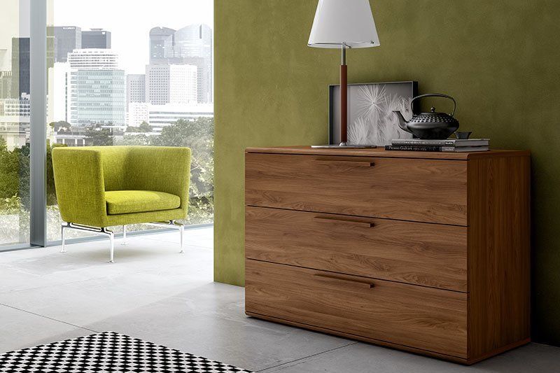 Soggiorno moderno con mobile in legno, lampada, sedia verde e vista sulla città attraverso ampie finestre.
