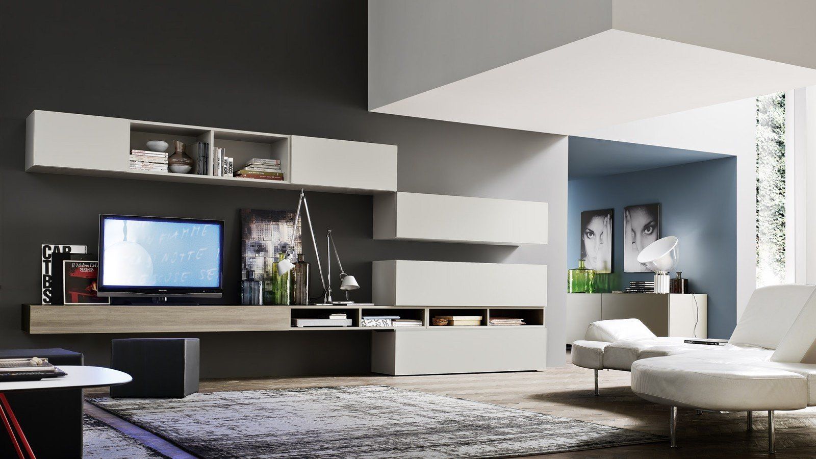 Soggiorno moderno con mensole a muro, TV, poltrone bianche, pouf e ampia finestra.