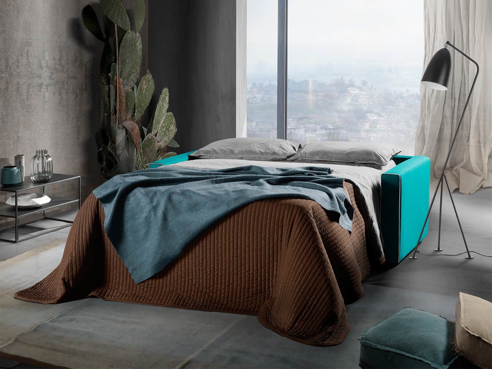 Camera da letto moderna con letto color verde acqua, lenzuola grigie, plaid marrone, ampia finestra e lampada da terra.