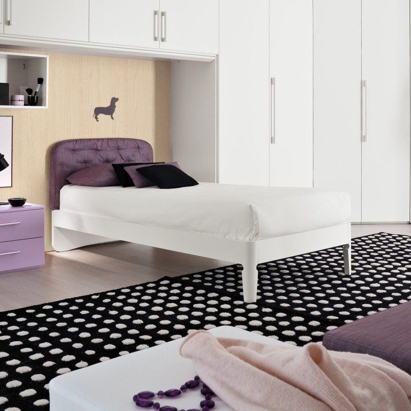 Camera da letto minimalista con letto bianco, dettagli viola, tappeto a pois neri e armadio bianco.