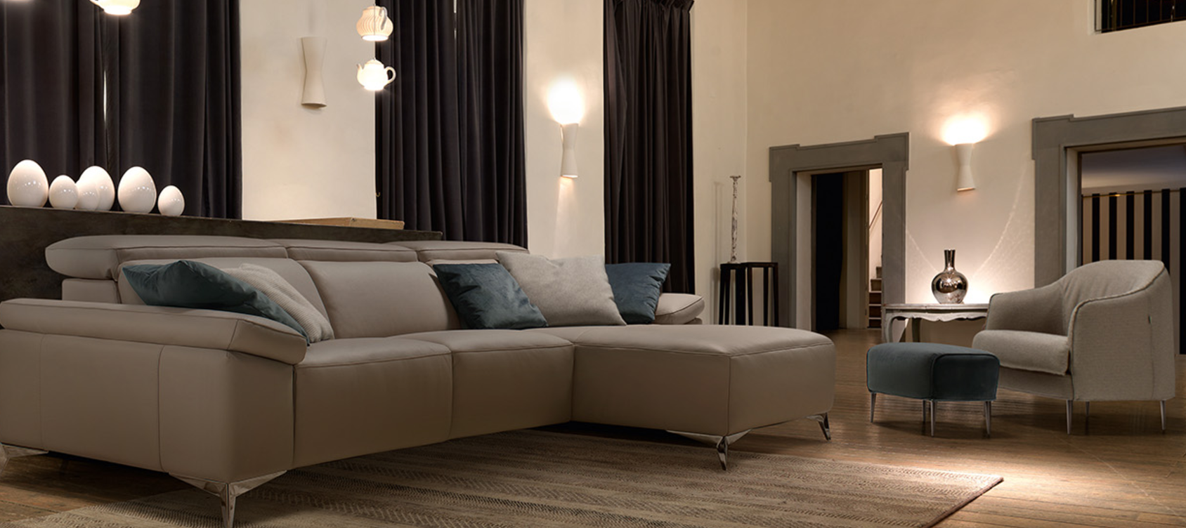 Soggiorno moderno con divano componibile beige, poltrona, lampade, tende e illuminazione soffusa e calda.