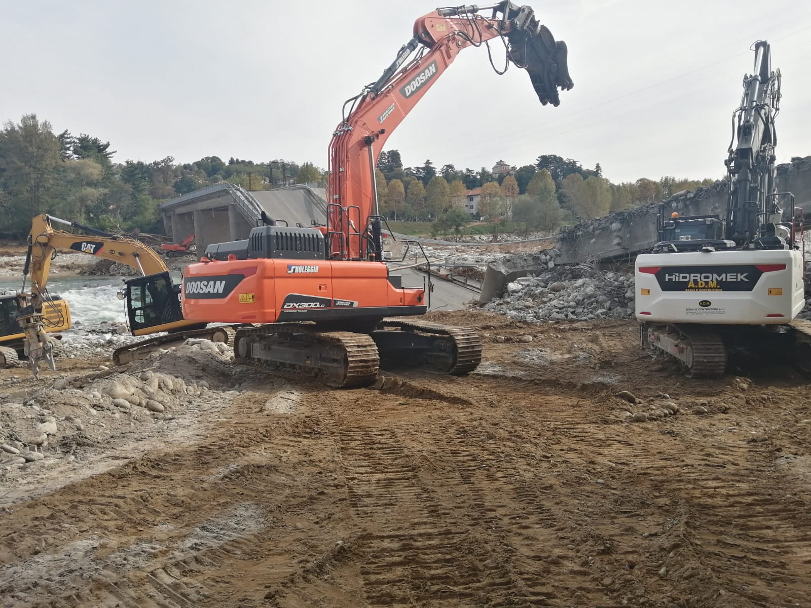 Tre escavatori pesanti lavorano su un terreno sterrato, demolendo detriti in un cantiere edile o di demolizione.