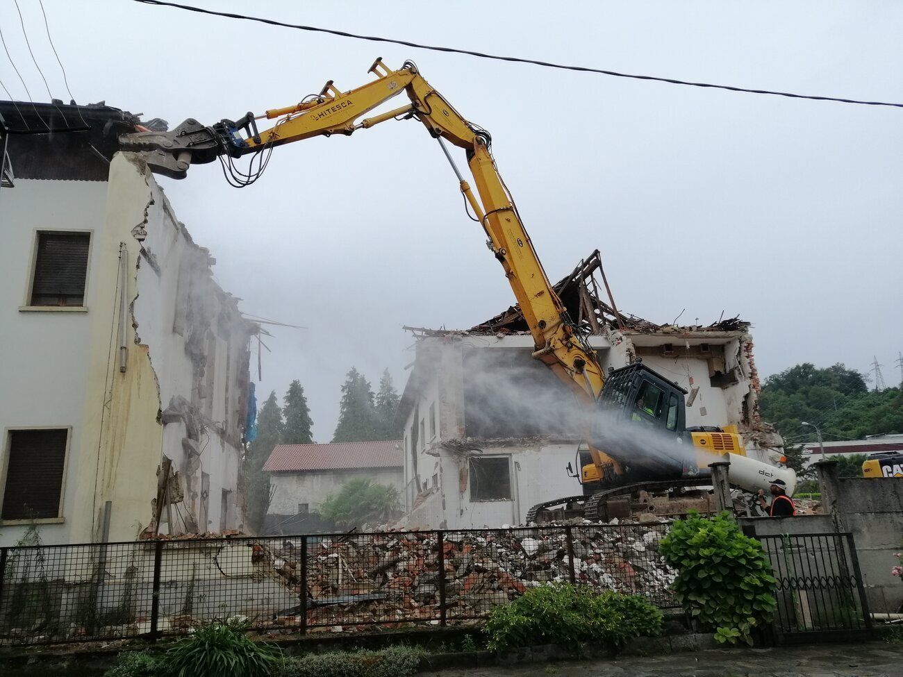 Un escavatore giallo da demolizione si protende verso il muro di un edificio, spruzzando acqua per sopprimere la polvere mentre abbatte i detriti.