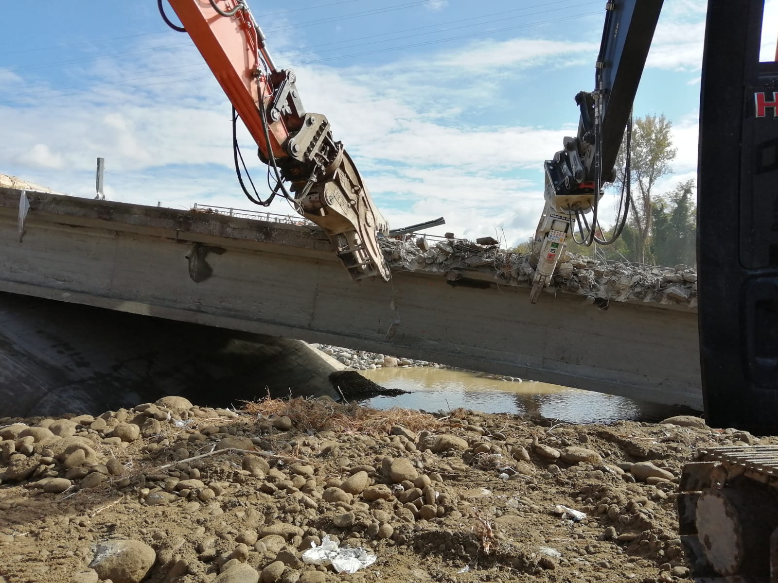 Sotto un cielo azzurro nuvoloso, due escavatori idraulici utilizzano frantumatori per calcestruzzo per demolire un ponte su un letto di fiume roccioso e asciutto.