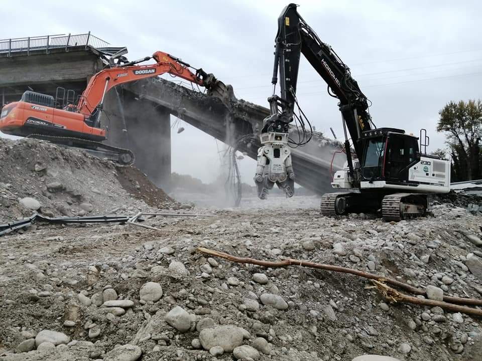 Due escavatori, uno arancione e uno nero, demoliscono la struttura di un ponte in cemento in un cantiere.